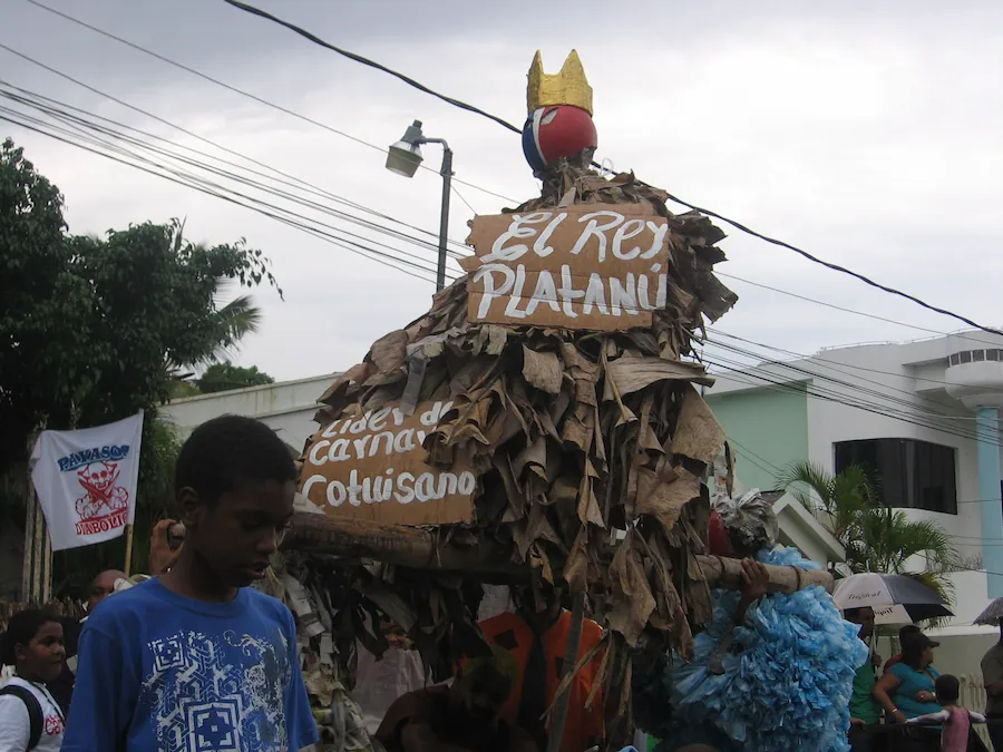Personaje del Rey Platanú con letreros artesanales siendo cargado en andas durante una celebración del carnaval tradicional de Cotuí.